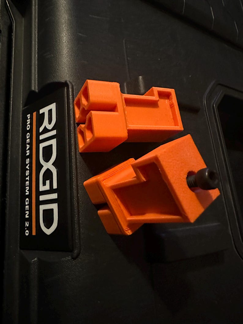 May include: Two bright orange, 3D-printed components with a rectangular design and interlocking features. One component has a black bolt. The items are on a black surface, near a label that reads "RIDGID PRO GEAR SYSTEM GEN 2.0".
