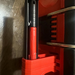 Op de afbeelding: Close-up van een rood en zwart gereedschap. Het gereedschap heeft een zwarte handgreep en een rode behuizing. Het gereedschap is gemaakt van plastic en heeft een geribbeld ontwerp. Het gereedschap is ontworpen om een ander gereedschap vast te houden. De achtergrond is een houten oppervlak.