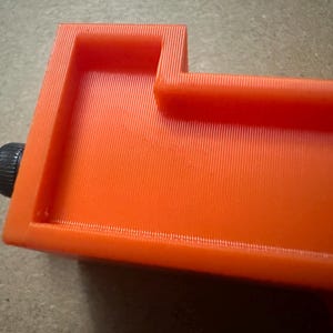 May include: Close-up of an orange 3D-printed object with a rectangular shape and a black knob. The object has a textured surface with vertical lines. The design includes raised edges and a recessed area.