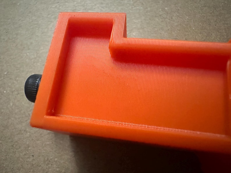 May include: Close-up of an orange 3D-printed object with a rectangular shape and a black knob. The object has a textured surface with vertical lines. The design includes raised edges and a recessed area.