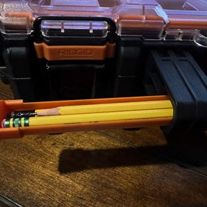 Puede incluir: Primer plano de una caja de herramientas RIDGID negra y naranja con un portal&aacute;pices. El portal&aacute;pices contiene tres l&aacute;pices: dos l&aacute;pices amarillos y un l&aacute;piz negro con una goma de borrar verde y plateada.