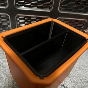 May include: An orange and black rectangular storage container with two compartments. The container has a textured exterior and a smooth, black interior. The background features a metallic, geometric pattern.