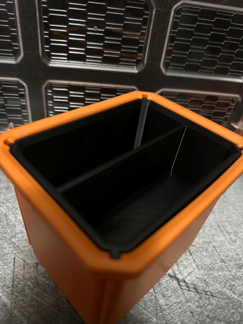 May include: An orange and black rectangular storage container with two compartments. The container has a textured exterior and a smooth, black interior. The background features a metallic, geometric pattern.