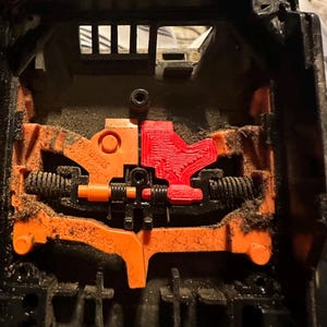 May include: Close-up of a black plastic object with orange and red internal components. The object has a complex mechanical design with springs and levers. The components are covered in dust.