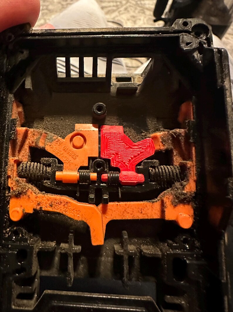 May include: Close-up of a black plastic object with orange and red internal components. The object has a complex mechanical design with springs and levers. The components are covered in dust.