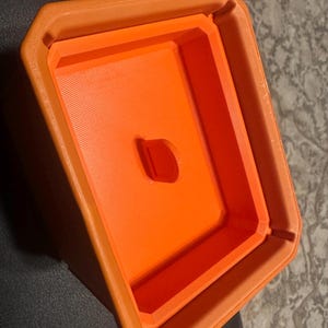May include: An orange, geometric-shaped container with a rectangular interior and a small, oval-shaped hole. The container has a textured surface and appears to be made of plastic. The design is simple and modern.