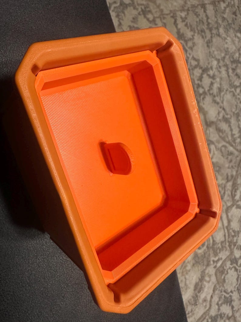 May include: An orange, geometric-shaped container with a rectangular interior and a small, oval-shaped hole. The container has a textured surface and appears to be made of plastic. The design is simple and modern.