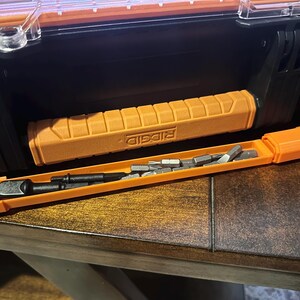 May include: An orange and black tool case with a matching orange handle and a separate orange bit holder. The bit holder contains several silver bits. The handle has the word "RIDGID" printed on it.