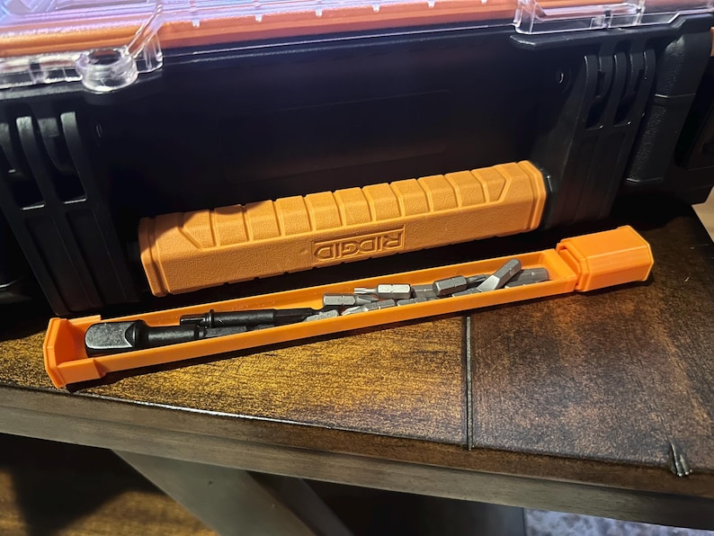 May include: An orange and black tool case with a matching orange handle and a separate orange bit holder. The bit holder contains several silver bits. The handle has the word "RIDGID" printed on it.