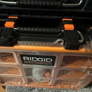 May include: A black and orange RIDGID Pro Gear System Gen 2.0 tool case. The case has a clear, orange-tinted front with a grid pattern. It features a black handle with orange accents and black latches.