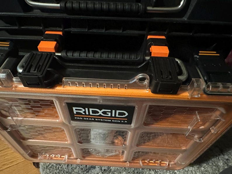 May include: A black and orange RIDGID Pro Gear System Gen 2.0 tool case. The case has a clear, orange-tinted front with a grid pattern. It features a black handle with orange accents and black latches.
