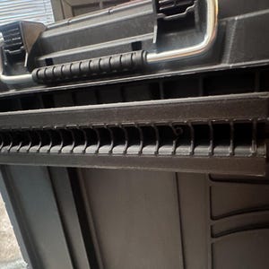 May include: Black plastic toolbox with a silver handle and latches. The toolbox has a textured surface and a series of slots along the front edge. The handle is made of metal. The toolbox is designed for storing tools and equipment.