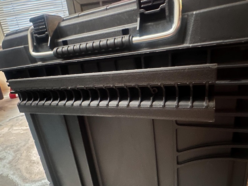 May include: Black plastic toolbox with a silver handle and latches. The toolbox has a textured surface and a series of slots along the front edge. The handle is made of metal. The toolbox is designed for storing tools and equipment.