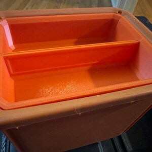 May include: An orange and brown plastic storage container with a rectangular shape. The container has a divider inside, creating two compartments. The interior is bright orange, while the exterior is a darker brown.