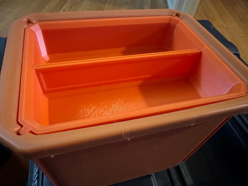 May include: An orange and brown plastic storage container with a rectangular shape. The container has a divider inside, creating two compartments. The interior is bright orange, while the exterior is a darker brown.
