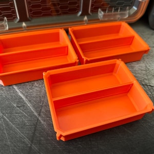 May include: Three bright orange storage trays with a divided interior are arranged on a textured gray surface. The trays are rectangular with rounded corners and a lip around the edge. A portion of a tool case is visible in the background.