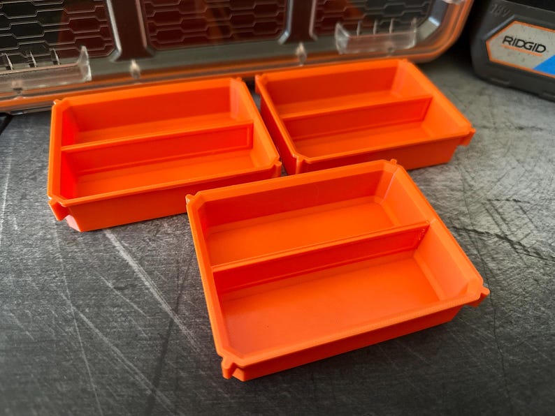 May include: Three bright orange storage trays with a divided interior are arranged on a textured gray surface. The trays are rectangular with rounded corners and a lip around the edge. A portion of a tool case is visible in the background.