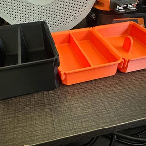 May include: Three plastic storage containers. One black container with two compartments and two orange containers with dividers. The orange containers have a handle. The containers are on a dark brown surface.