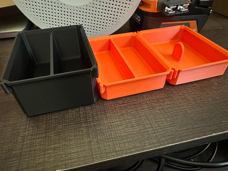 May include: Three plastic storage containers. One black container with two compartments and two orange containers with dividers. The orange containers have a handle. The containers are on a dark brown surface.