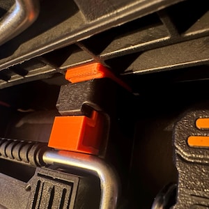 May include: Close-up of a black tool case interior with orange and silver accents. The case features several clamps and supports, with bright orange locking mechanisms. The design suggests a focus on organization and secure storage for tools.