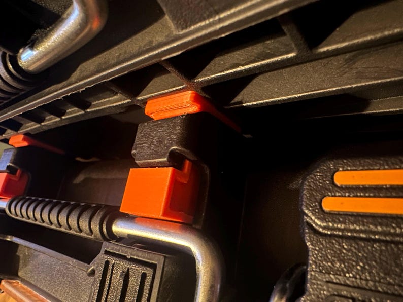 May include: Close-up of a black tool case interior with orange and silver accents. The case features several clamps and supports, with bright orange locking mechanisms. The design suggests a focus on organization and secure storage for tools.