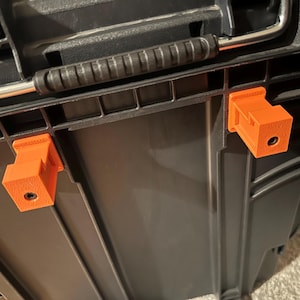 May include: Close-up of a black storage container with an orange 3D-printed clip attached. The clip is rectangular with a hole and is mounted on the side of the container. The container has a black handle and a silver metal bar.
