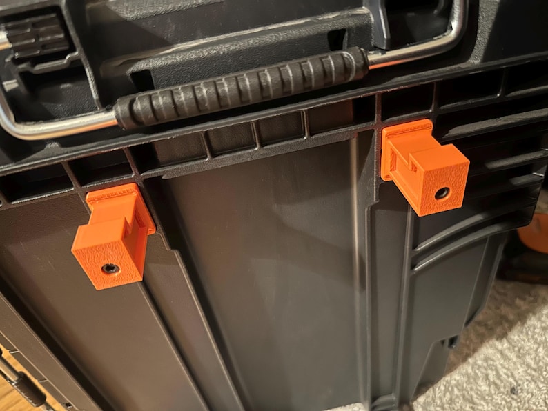 May include: Close-up of a black storage container with an orange 3D-printed clip attached. The clip is rectangular with a hole and is mounted on the side of the container. The container has a black handle and a silver metal bar.