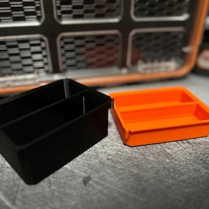 May include: Two small plastic storage trays. One is black with two compartments, and the other is bright orange with two compartments. The trays are designed to fit inside a larger case, with a textured background.