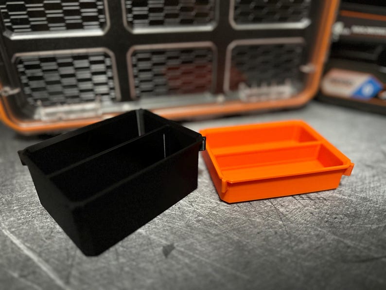 May include: Two small plastic storage trays. One is black with two compartments, and the other is bright orange with two compartments. The trays are designed to fit inside a larger case, with a textured background.