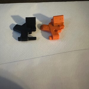 May include: Two plastic toy figures, one black and one orange, are displayed on a white surface. The figures have a similar design, with a rectangular head and a body with a curved section and a small foot.