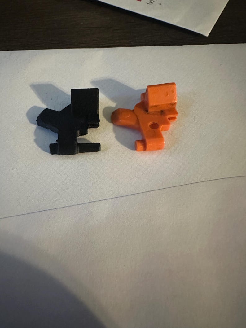 May include: Two plastic toy figures, one black and one orange, are displayed on a white surface. The figures have a similar design, with a rectangular head and a body with a curved section and a small foot.