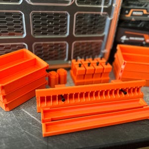 Puede incluir: Un surtido de contenedores y organizadores de almacenamiento de color naranja brillante. La imagen muestra varios contenedores rectangulares, soportes cilíndricos y un organizador tipo rack, todos en un vibrante color naranja. Los artículos parecen ser de plástico y están diseñados para guardar herramientas.