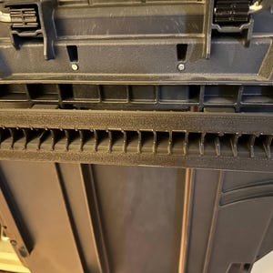 May include: Close-up of a black plastic toolbox with a textured surface. The toolbox has a series of rectangular slots and a hinged lid with black latches. The interior of the toolbox is visible.