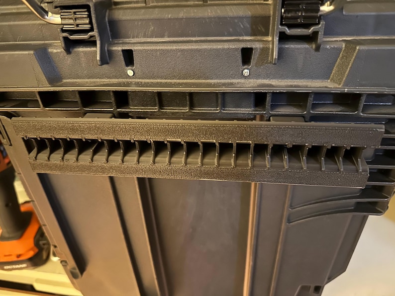 May include: Close-up of a black plastic toolbox with a textured surface. The toolbox has a series of rectangular slots and a hinged lid with black latches. The interior of the toolbox is visible.