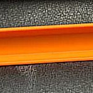May include: An orange plastic rectangular tube with a hollow interior. The tube has a rounded end and a small lip on one side. The object is set against a dark, textured surface.
