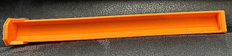May include: An orange plastic rectangular tube with a hollow interior. The tube has a rounded end and a small lip on one side. The object is set against a dark, textured surface.