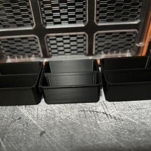 May include: Black plastic storage containers with dividers, arranged in a row. The containers are designed to fit inside a larger orange and black storage case. The case has a grid-like pattern on the lid.