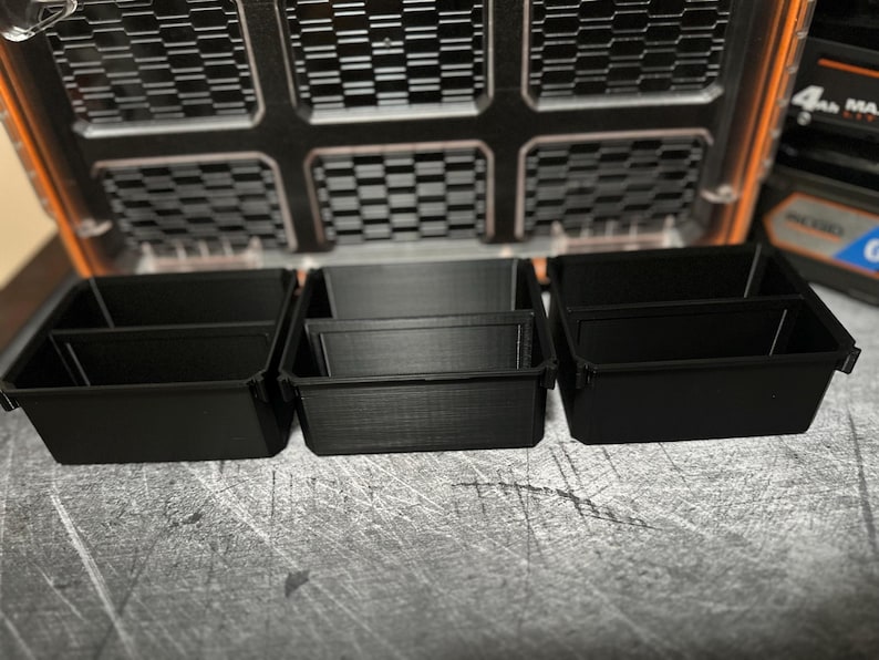 May include: Black plastic storage containers with dividers, arranged in a row. The containers are designed to fit inside a larger orange and black storage case. The case has a grid-like pattern on the lid.