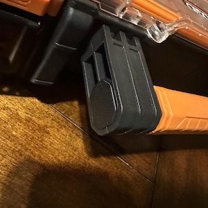 May include: Close-up of a black and orange tool box latch. The latch is made of black plastic and is attached to an orange handle. The tool box has a clear top and is labeled with the brand name "RIDGID".