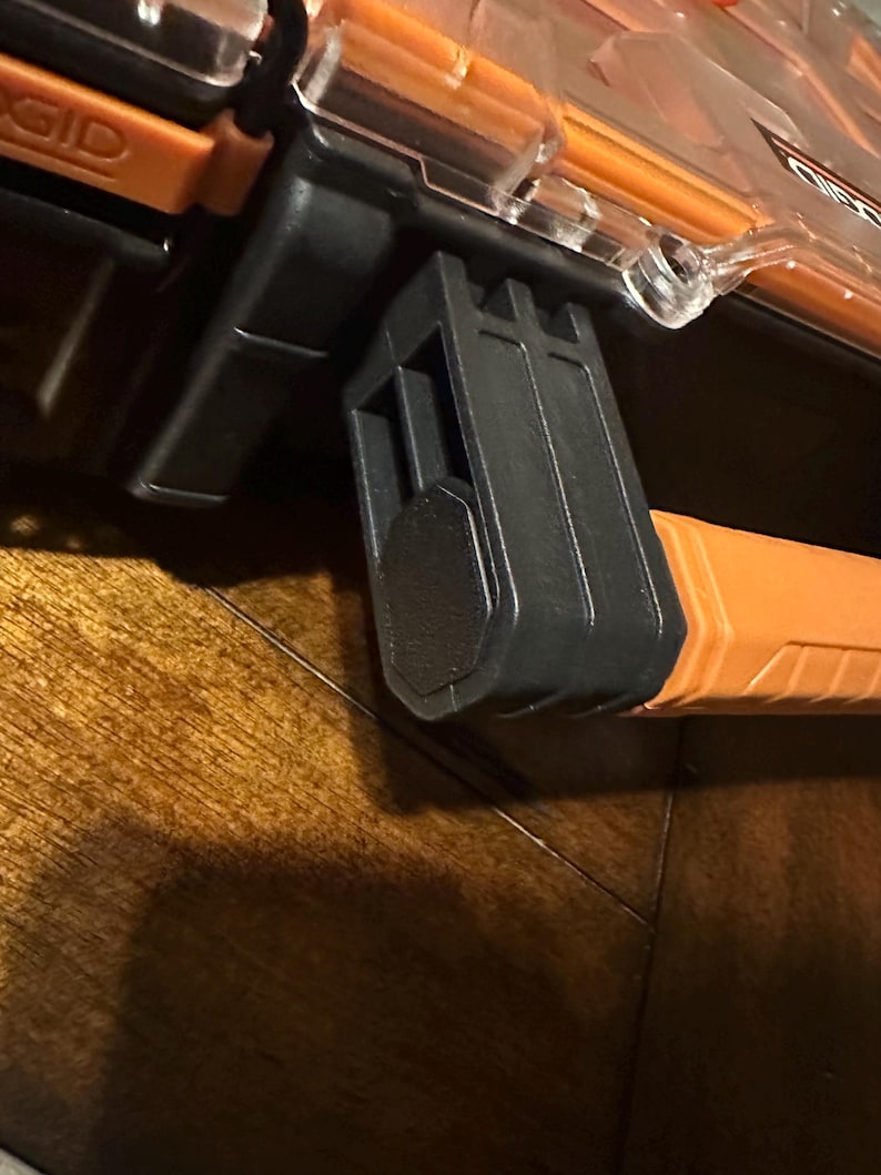 May include: Close-up of a black and orange tool box latch. The latch is made of black plastic and is attached to an orange handle. The tool box has a clear top and is labeled with the brand name "RIDGID".