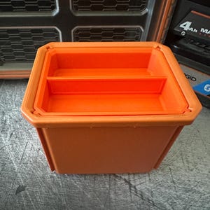 May include: An orange plastic storage container with a divided interior. The container is rectangular with a slightly tapered design. The container is sitting on a gray surface. Other items are visible in the background.