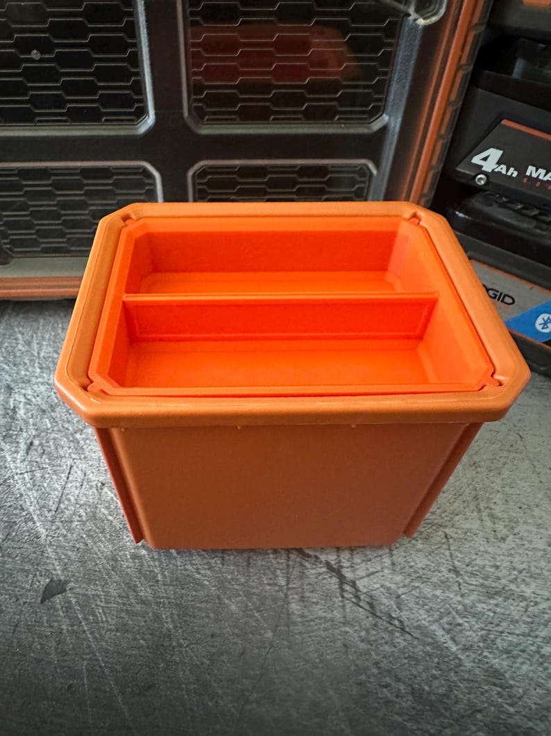 May include: An orange plastic storage container with a divided interior. The container is rectangular with a slightly tapered design. The container is sitting on a gray surface. Other items are visible in the background.