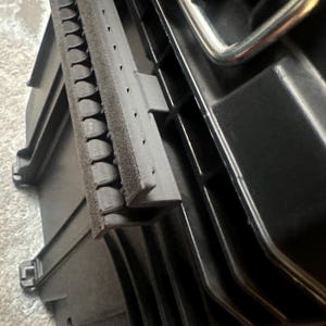 May include: Black plastic storage case with a silver handle. The case has a textured surface and a foam insert. The lid is open, revealing the interior. The case is designed for tools or equipment.