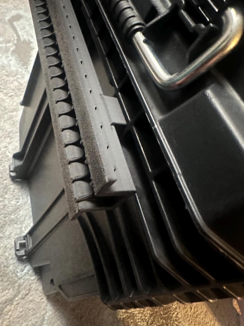 May include: Black plastic storage case with a silver handle. The case has a textured surface and a foam insert. The lid is open, revealing the interior. The case is designed for tools or equipment.