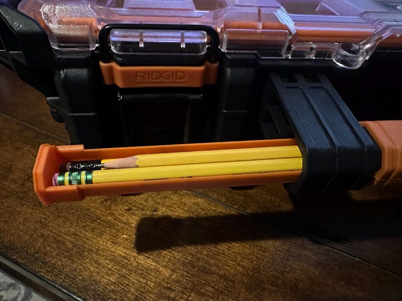 May include: A black and orange RIDGID tool box with a clear lid. Three yellow pencils and one black pencil are stored in an orange compartment. The pencils are visible in the foreground.