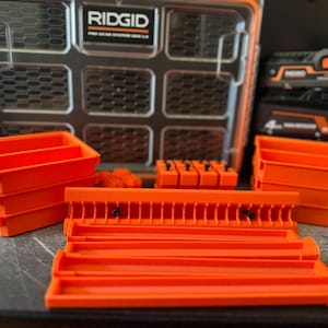 May include: An orange and black RIDGID Pro Gear System 2.0 tool organizer with various orange storage containers and accessories. The case is open, revealing multiple compartments. The image also shows power tool batteries.