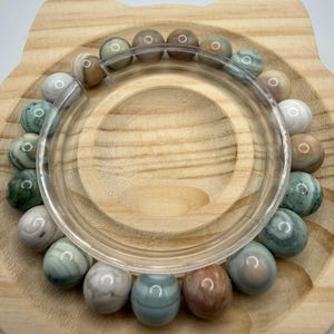 May include: A beaded bracelet featuring smooth, round stones in various shades of green, brown, white, and grey. The beads are arranged in a circular pattern, showcasing natural color variations and textures within each individual stone bead.