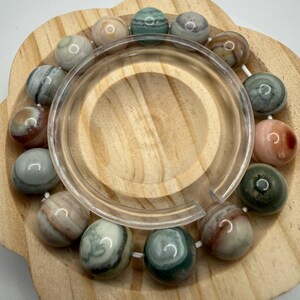 May include: A circular bracelet composed of smooth, polished spherical beads in various earthy tones including shades of green, brown, cream, and grey. The beads are strung together on a white cord, displaying natural marbling and color variations.