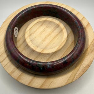 May include: A round, burgundy-colored bangle bracelet with flecks of green and black. The bracelet is resting on a light-colored wooden plate. A small white sticker with the number "57" is affixed to the bracelet.