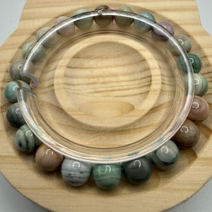 May include: A circular bracelet composed of smooth, multi-colored stone beads in shades of sage green, dusty pink, grey, and cream. The beads feature natural banded patterns and are arranged in a continuous loop, resting on a light wood display.